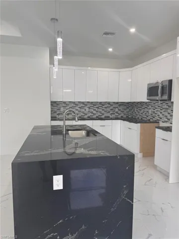 Kitchen featuring dark stone counters, a kitchen island with sink, sink, decorative light fixtures, and white cabinets