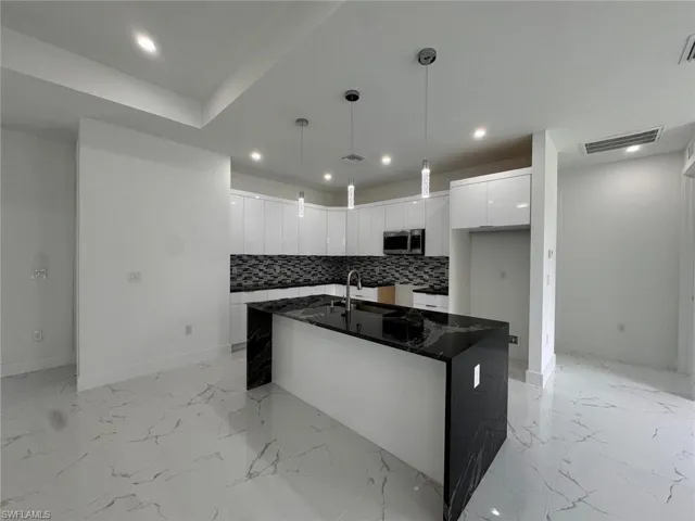 Kitchen with a kitchen island with sink, dark stone counters, decorative backsplash, decorative light fixtures, and white cabinetry