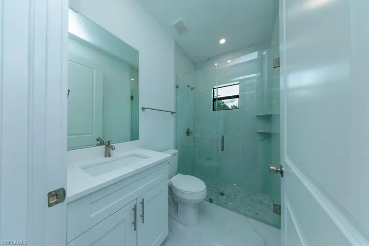 Bathroom featuring a white vanity with integrated sink, brushed nickel faucet, large mirror, and glass-enclosed shower with tiled walls and pebble-finish floor