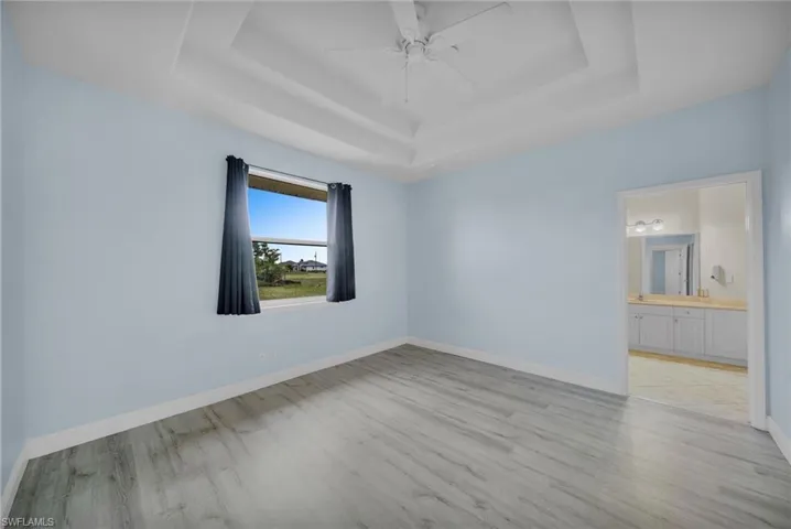 Unfurnished bedroom with a tray ceiling, a ceiling fan, light wood-type flooring, and connected bathroom