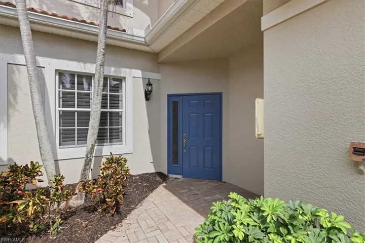 Inviting entryway featuring a blue front door with a sidelight, a brick paver walkway, and lush landscaping