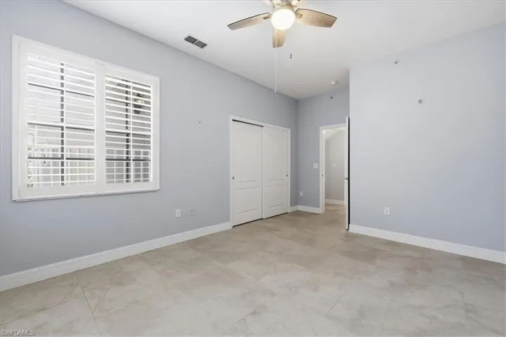 Spacious room featuring light-toned tile flooring, a window with plantation shutters, dual bifold closet doors, and a ceiling fan with integrated lighting