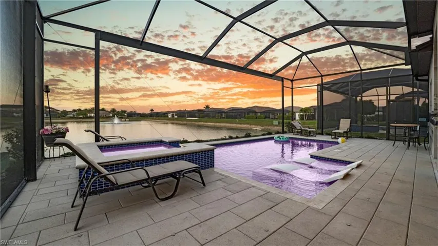 Pool at dusk with a sunroom, glass enclosure, a water view, a patio, and a pool with connected hot tub