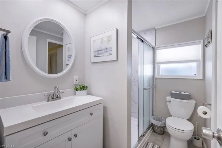 Well-appointed bathroom featuring a white vanity with a rectangular sink, a circular mirror, a shower with a sliding glass door, and a window providing natural light