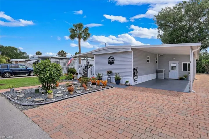 The property features a brick paver driveway, a covered carport, and a decorative garden bed with various plants and stones