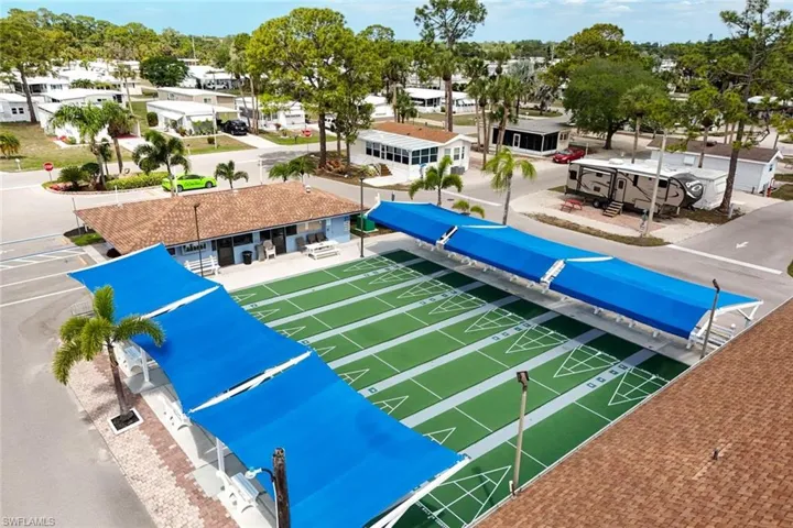 The property features a well-maintained shuffleboard court with blue overhead canopies providing shade, bordered by a paved area and various trees
