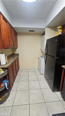 Kitchen with light tile patterned floors, stacked washer and clothes dryer, dark stone counters, and fridge