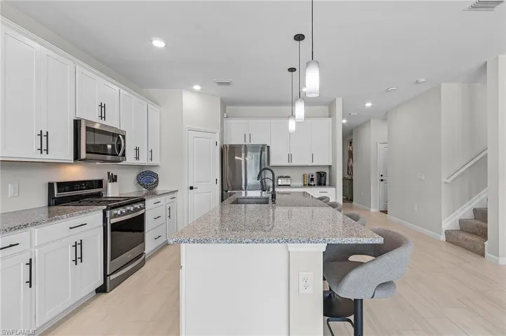 Contemporary kitchen featuring an oversized island with pendant lighting, stone countertops, white cabinetry with matte black hardware, stainless steel appliances, and wood-finish flooring