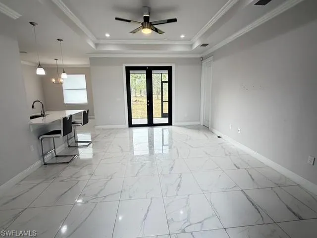 Unfurnished living room featuring a raised ceiling, french doors, light marble finish floors, crown molding, and a ceiling fan