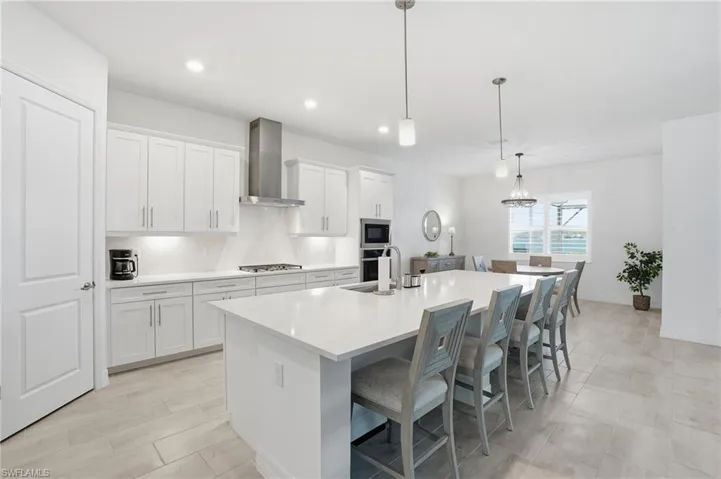 Kitchen featuring pendant lighting, a kitchen breakfast bar, wall chimney exhaust hood, a center island with sink, and white cabinets