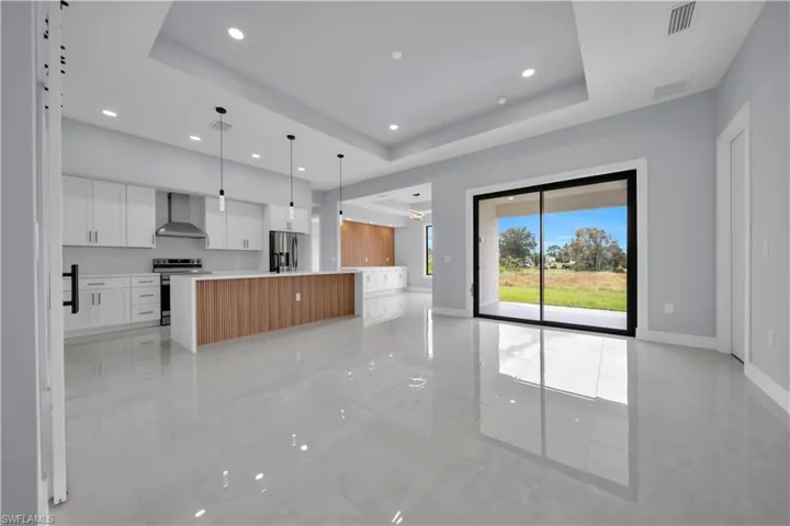 Open-concept living space with large format tile flooring, a kitchen featuring white cabinetry and stainless steel appliances, a kitchen island with wood-finish paneling, and a tray ceiling with recessed lighting