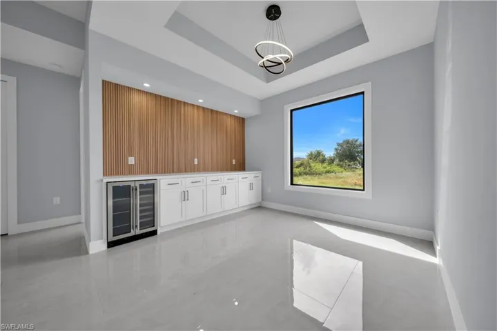 Tray ceiling with a contemporary chandelier, large picture window, built-in cabinetry, dual beverage refrigerators, and wood-panel accent wall