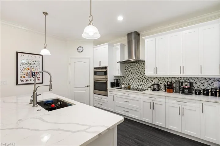 Kitchen featuring  chimney exhaust hood, backsplash, white cabinetry, and hanging light fixtures