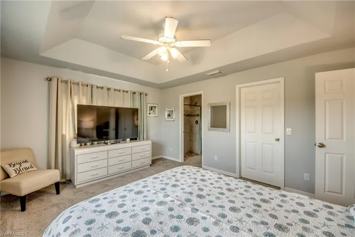 Tray ceiling with a ceiling fan, light gray wall paint, light-colored carpeting, and a visible doorway to a shower area
