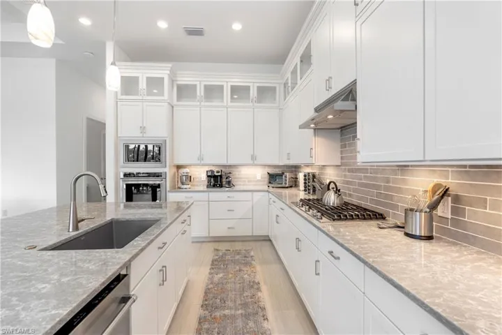 Kitchen featuring glass insert cabinets, under cabinet range hood, white cabinetry, light stone counters, and hanging light fixtures