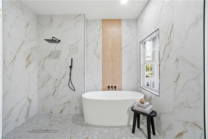 A spa-inspired primary wet room featuring large-format marble-look porcelain tile, a sleek walk-in shower, freestanding soaking tub, matte black fixtures, and beautiful natural light.