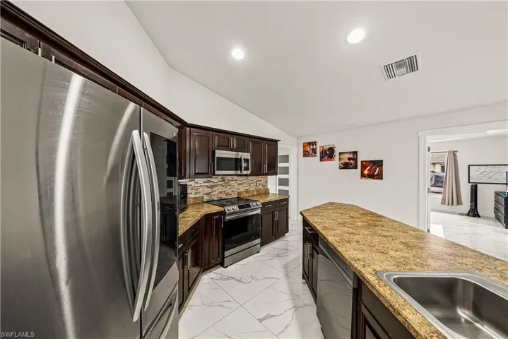 Kitchen with stainless steel appliances, dark wood finish cabinets, lofted ceiling, light marble finish flooring, and decorative backsplash