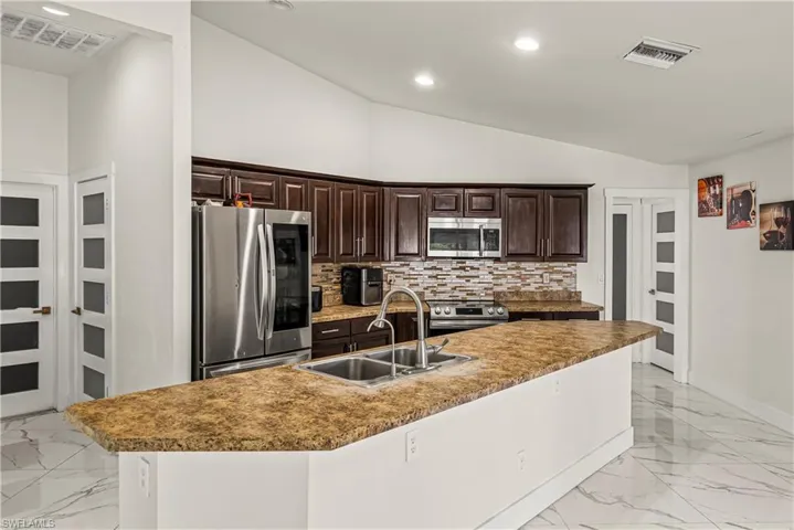 Kitchen featuring dark wood finish cabinetry, lofted ceiling, stainless steel appliances, light marble finish floors, and decorative backsplash