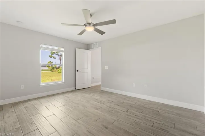 Spacious room featuring wood-finish flooring, a contemporary ceiling fan with integrated lighting, a large window providing natural illumination, a white paneled door, and neutral wall tones