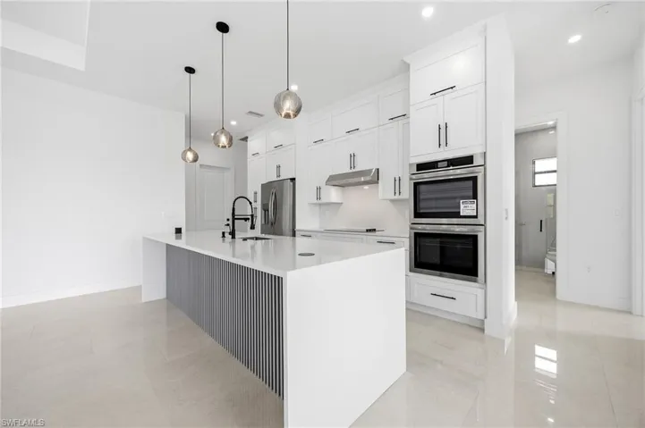 Kitchen with white cabinets, stainless steel appliances, light stone countertops, decorative light fixtures, and an island with sink