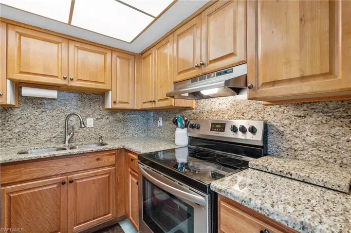 Kitchen with decorative backsplash, under cabinet range hood, a sink, light stone counters, and stainless steel electric stove