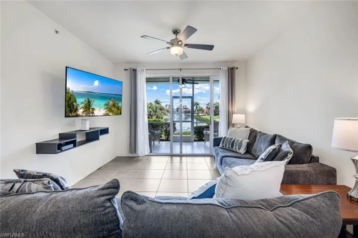 Living area with sliding glass doors opening to a water view, featuring a ceiling fan with integrated lighting, a wall-mounted media console, and tile flooring