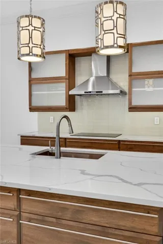 Kitchen with brown cabinetry, wall chimney range hood, pendant lighting, ornamental molding, and backsplash