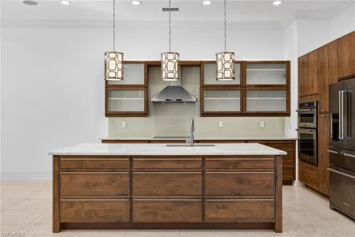 Kitchen with tasteful backsplash, an island with sink, glass insert cabinets, light stone counters, and ornamental molding