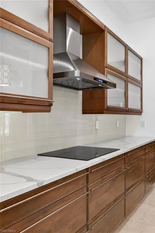 Kitchen featuring brown cabinets, wall chimney range hood, backsplash, light stone counters, and glass insert cabinets