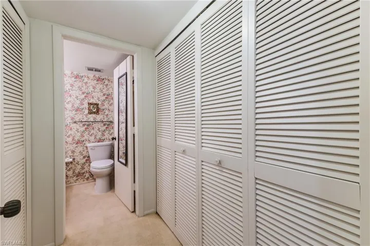 Hallway with multiple louvered closet doors, leading to a room featuring floral wallpaper and a toilet