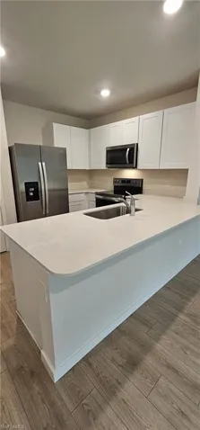 Kitchen featuring a large island with an undermount sink, light-toned countertops, white cabinetry, stainless steel appliances, and wood-finish flooring