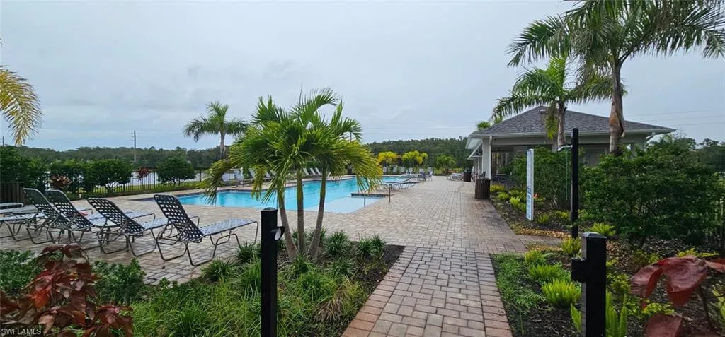 Expansive paver patio surrounding a large swimming pool, complemented by mature palm trees, lush landscaping, and a distant water feature