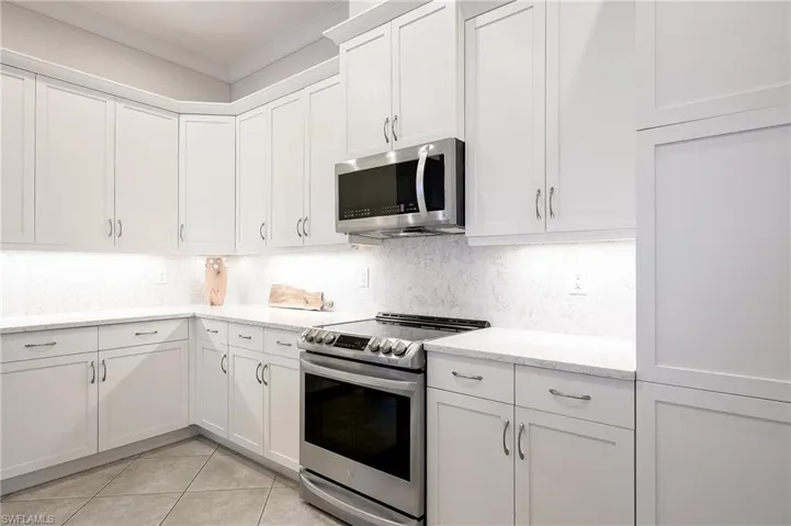 Kitchen with light tile patterned floors, stainless steel appliances, ornamental molding, light stone counters, and white cabinets