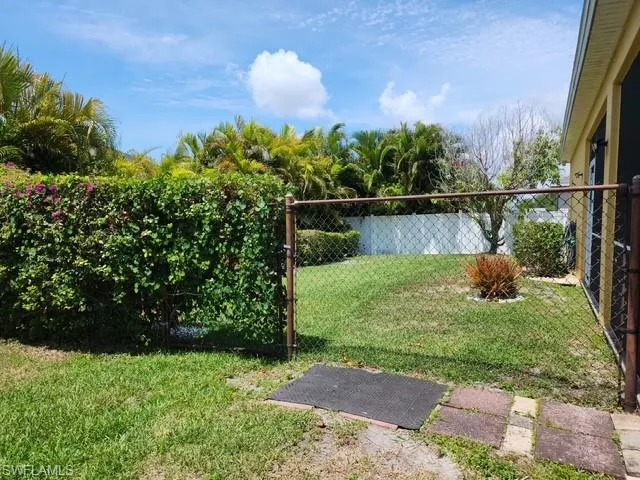 Enclosed backyard featuring a chain-link fence, a lush green hedge, mature palm trees, and a grassy area with a small decorative plant