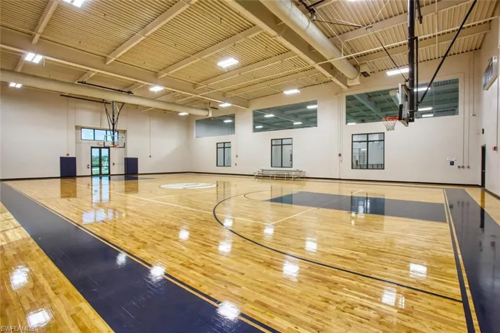 Full-size indoor basketball court featuring polished wood-finish flooring, high-clearance exposed industrial ceiling, integrated adjustable basketball hoops, and perimeter windows