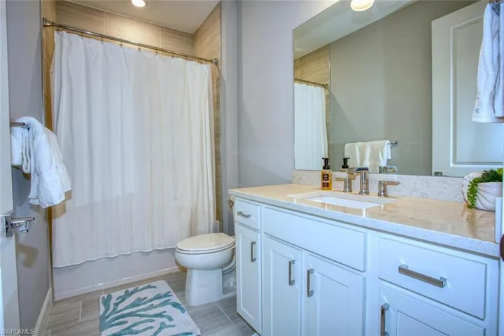 Bathroom featuring white shaker-style cabinetry, stone-finish countertop with an integrated rectangular sink, large wall-mounted mirror, and tile surround within the tub area
