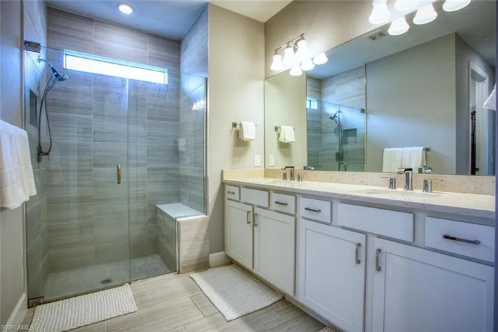 Bathroom featuring white cabinetry, quartz countertop with dual undermount sinks, full-width mirror, and a walk-in shower with floor-to-ceiling tile, glass enclosure, and built-in bench