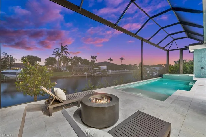 Pool at dusk featuring a patio area, a water view, an outdoor fire pit, a lanai, and a pool with connected hot tub