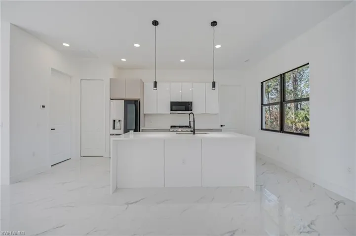 Kitchen featuring decorative light fixtures, an island with sink, stainless steel fridge, light stone counters, and white cabinets