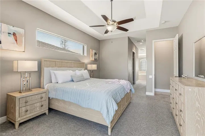 Bedroom with ensuite bathroom, light colored carpet, ceiling fan, and a tray ceiling