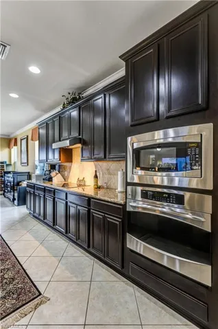 Kitchen with backsplash, appliances with stainless steel finishes, light stone counters, dark cabinets, and light tile patterned floors