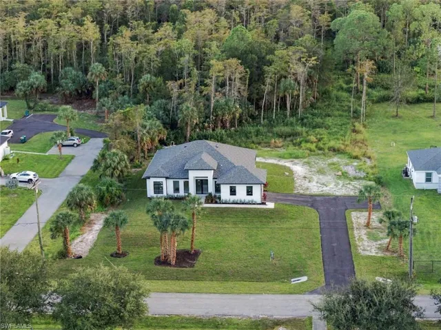Single-story residence featuring a hipped shingle roof, white stucco exterior, asphalt driveway, and landscaped lawn with mature palm trees, bordered by native woodlands
