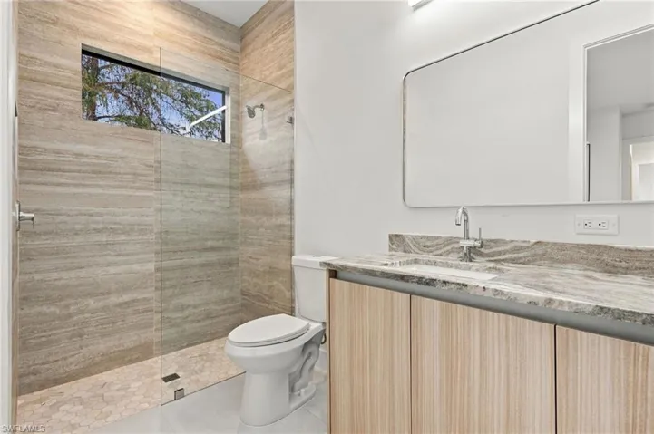 Bathroom featuring a stone-tile walk-in shower with a glass panel, wood-finish cabinetry, natural stone countertop with an integrated sink, and a large-format rectangular wall mirror