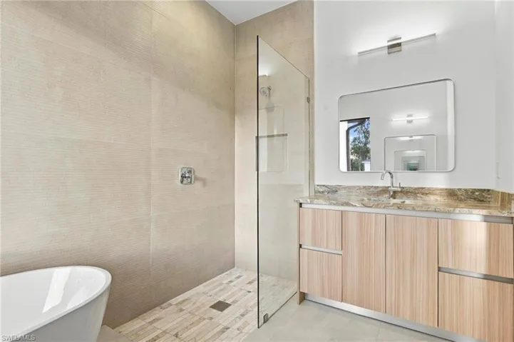 Bathroom featuring a freestanding tub, walk-in shower with a frameless glass enclosure, wood-finish vanity cabinetry, stone-patterned countertop, and a wall-mounted mirror