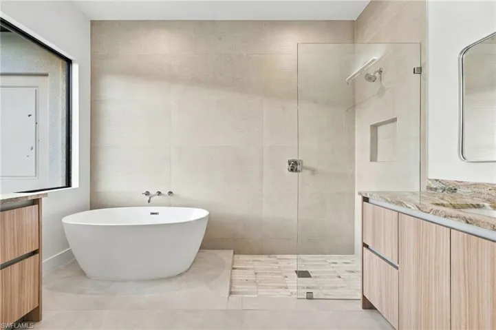 Freestanding white soaking tub with wall-mounted faucet, walk-in glass-enclosed shower, built-in shower niche, stone-finish vanity countertops, and wood-finish cabinetry