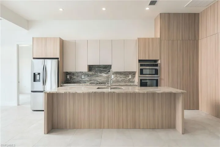 Kitchen featuring wood-finish cabinetry, stone slab backsplash, expansive stone-topped island, stainless steel wall-mounted oven and microwave, and built-in refrigerator