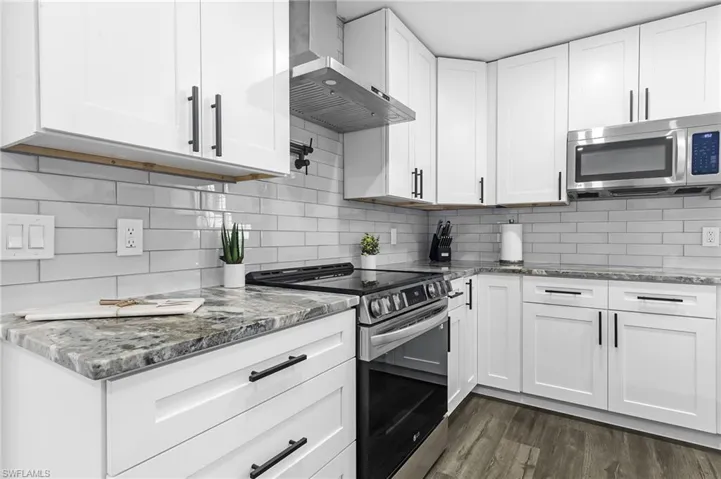 Kitchen featuring wall chimney range hood, white cabinets, light stone countertops, appliances with stainless steel finishes, and dark wood-style floors