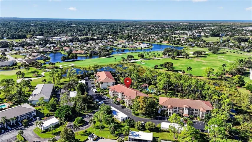 Aerial perspective of a multi-story residential complex featuring terracotta-tiled rooflines, paved surface parking, and mature landscaping, situated within a setting of expansive green space and water features