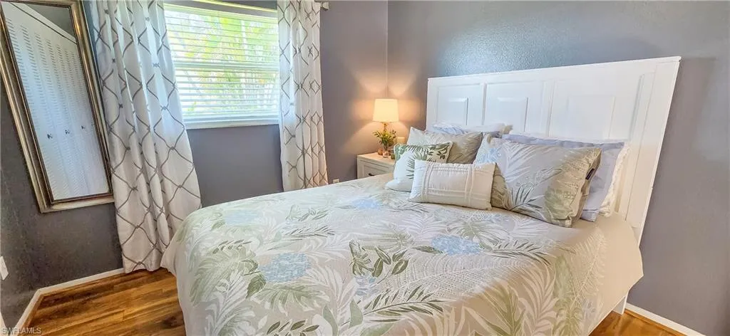 Bedroom featuring wood-finish flooring, a white paneled headboard, and a window with white blinds and decorative curtains