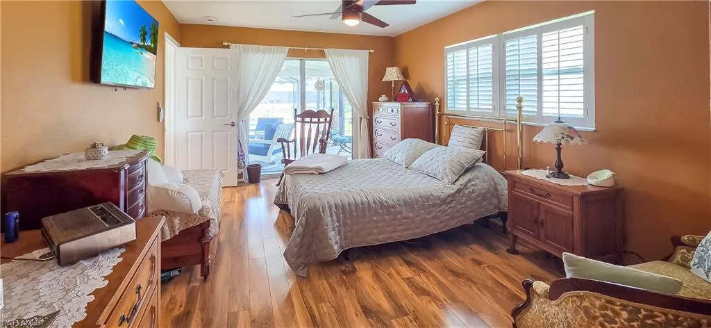 Wood-finish flooring throughout, featuring a ceiling fan, sliding glass doors with white drapes, and two windows with white louvered shutters